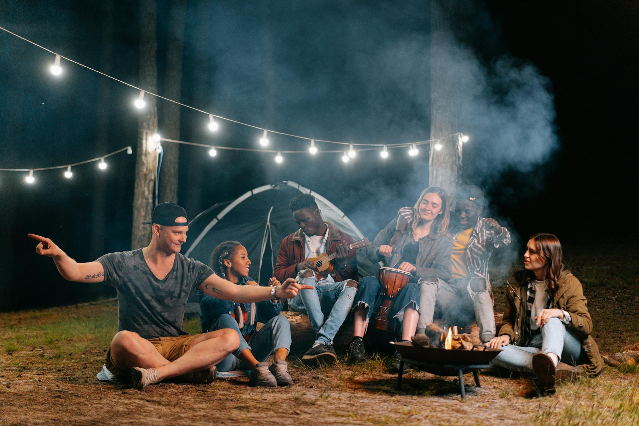 A group of friends relaxes by a campfire in the forest, playing music and talking.